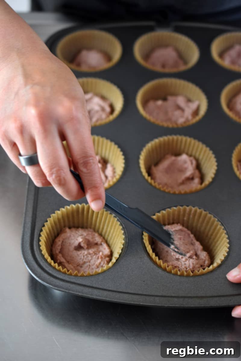 A hand uses a small, sleek black spatula to meticulously smooth out the pink strawberry muffin batter within a parchment-lined muffin pan, creating an even bottom layer.