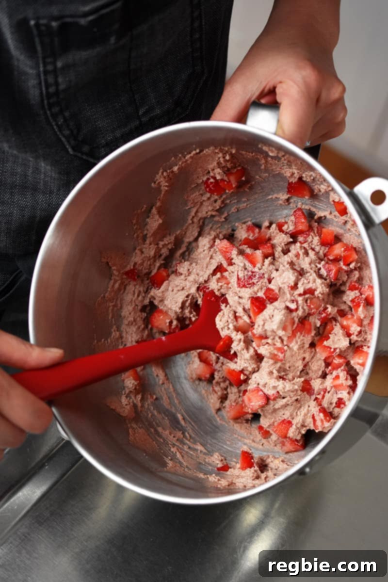 An overhead view shows a hand using a red spatula to gently fold diced fresh strawberries into the remaining vibrant pink muffin batter, ensuring even distribution.