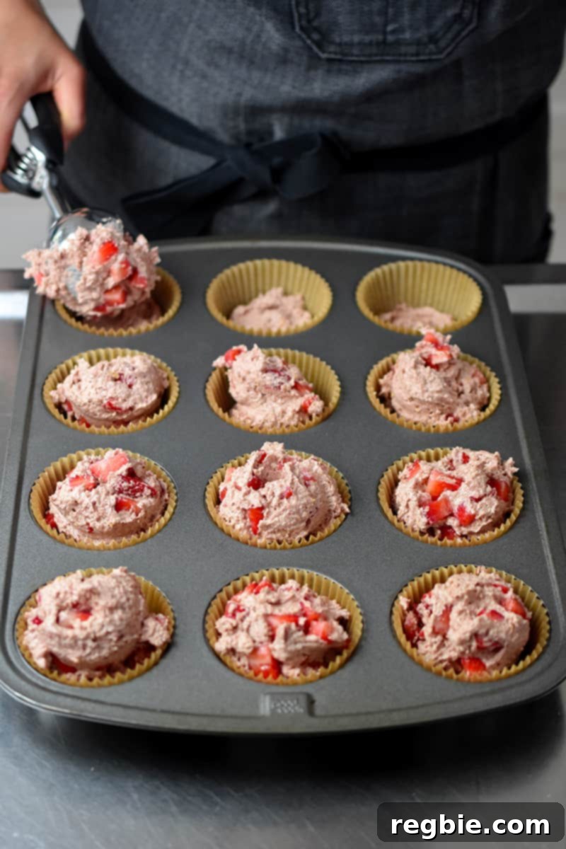 The process of adding scoops of strawberry muffin batter to a muffin tin, which already contains a small, fruit-free layer of batter at the bottom of each parchment liner.