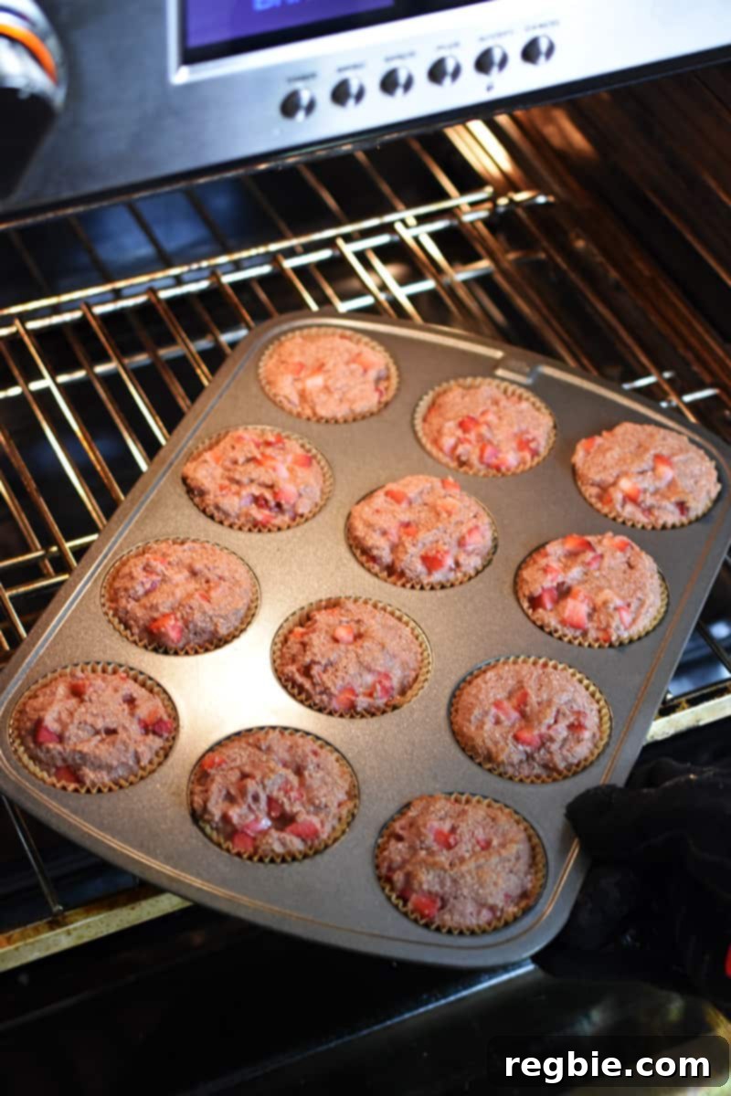 Someone reaches into an open oven to rotate a muffin pan, filled with beautifully rising strawberry muffins, indicating the halfway point of their baking process.