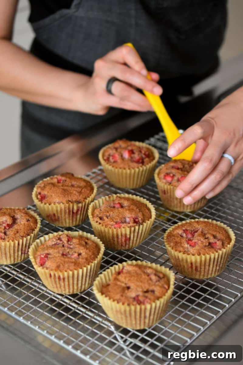 Two hands are carefully transferring freshly baked gluten-free paleo strawberry muffins from the muffin pan onto a metal cooling rack to cool down.