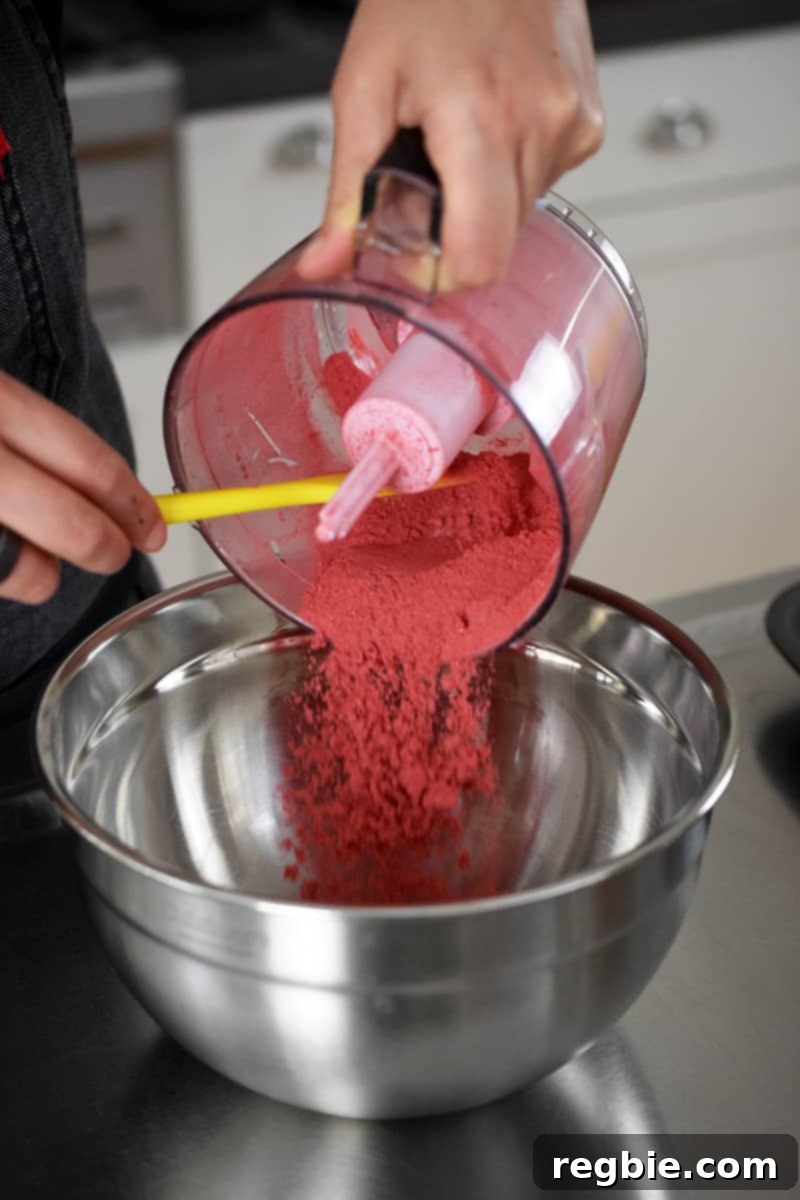 An individual carefully pouring powdered freeze-dried strawberries, freshly blitzed from a small food processor, into a larger silver mixing bowl, ready to be incorporated into the muffin batter.