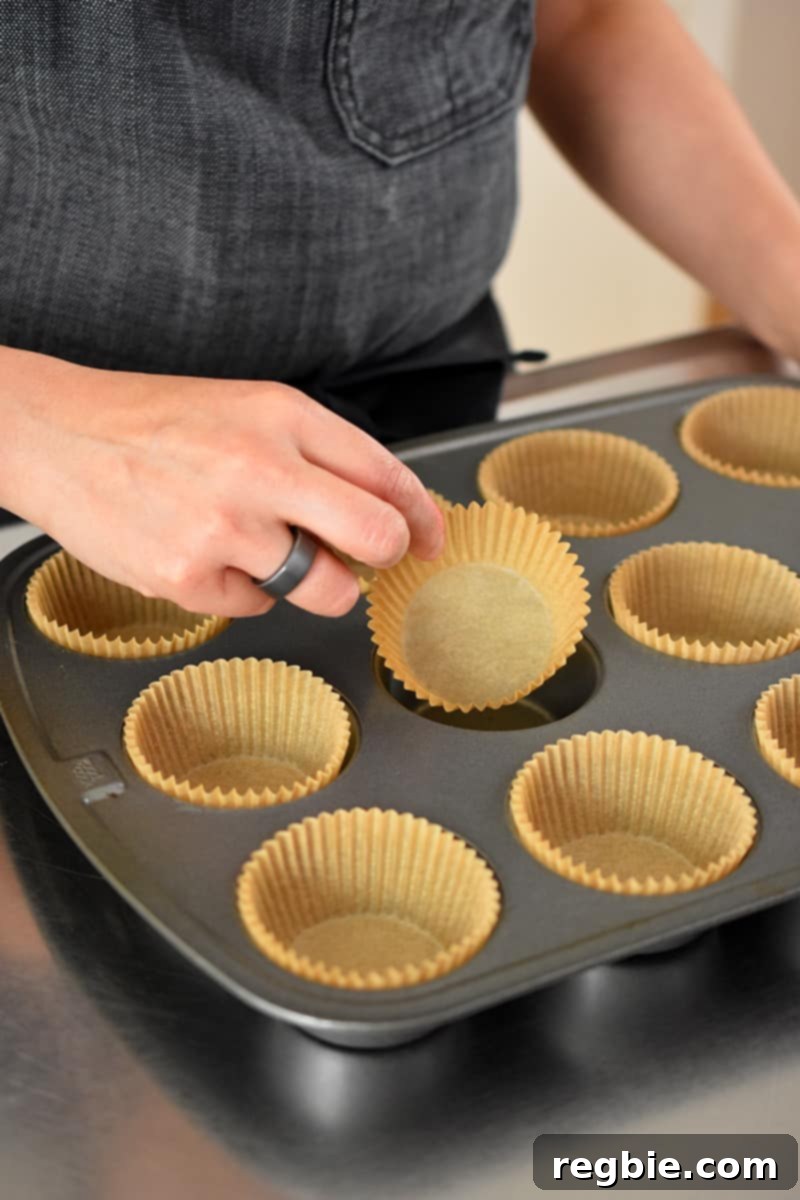 A hand gently places a crinkled parchment muffin liner into a compartment of a standard muffin tin, preparing it for the batter.
