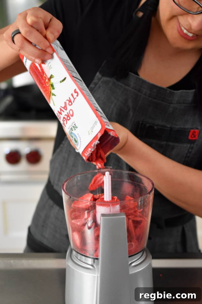 A woman with a warm smile is seen carefully pouring a package of vibrant freeze-dried strawberries into the bowl of a small food processor, ready for processing.