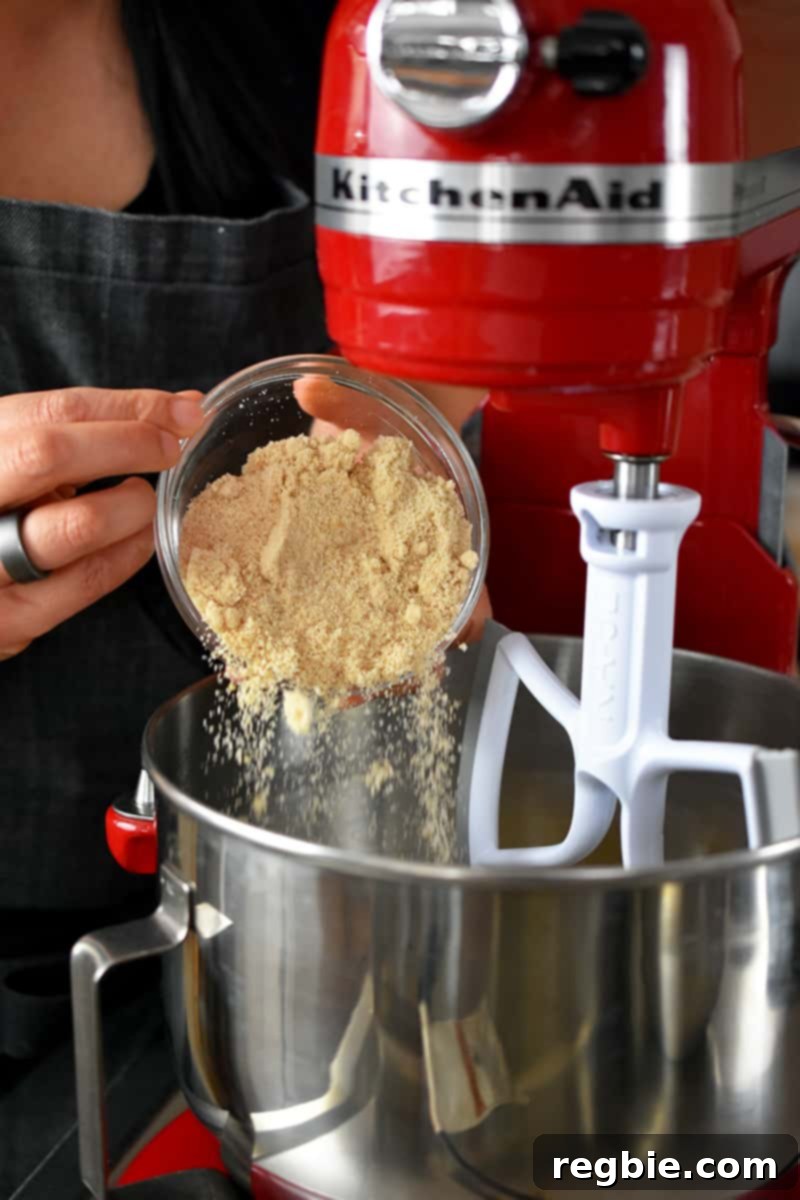 Someone in a gray apron gently pours a small bowl of maple sugar into the shiny silver bowl of a bright red stand mixer, ready to be creamed with the ghee.