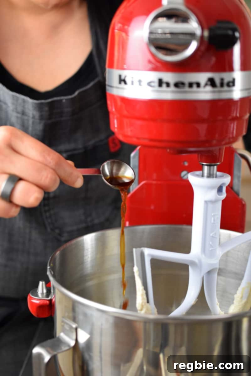 Someone wearing a gray apron is shown adding a spoonful of clear vanilla extract into the mixing bowl of a red Kitchenaid stand mixer, alongside other wet ingredients.