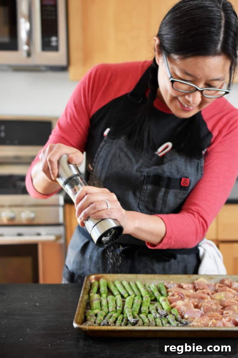 Simple Sheet Pan Chicken & Asparagus: Paleo Perfection 12 An Asian woman is adding freshly ground black pepper from a pepper mill onto a bed of green asparagus spears arranged on a sheet pan, ready for roasting.