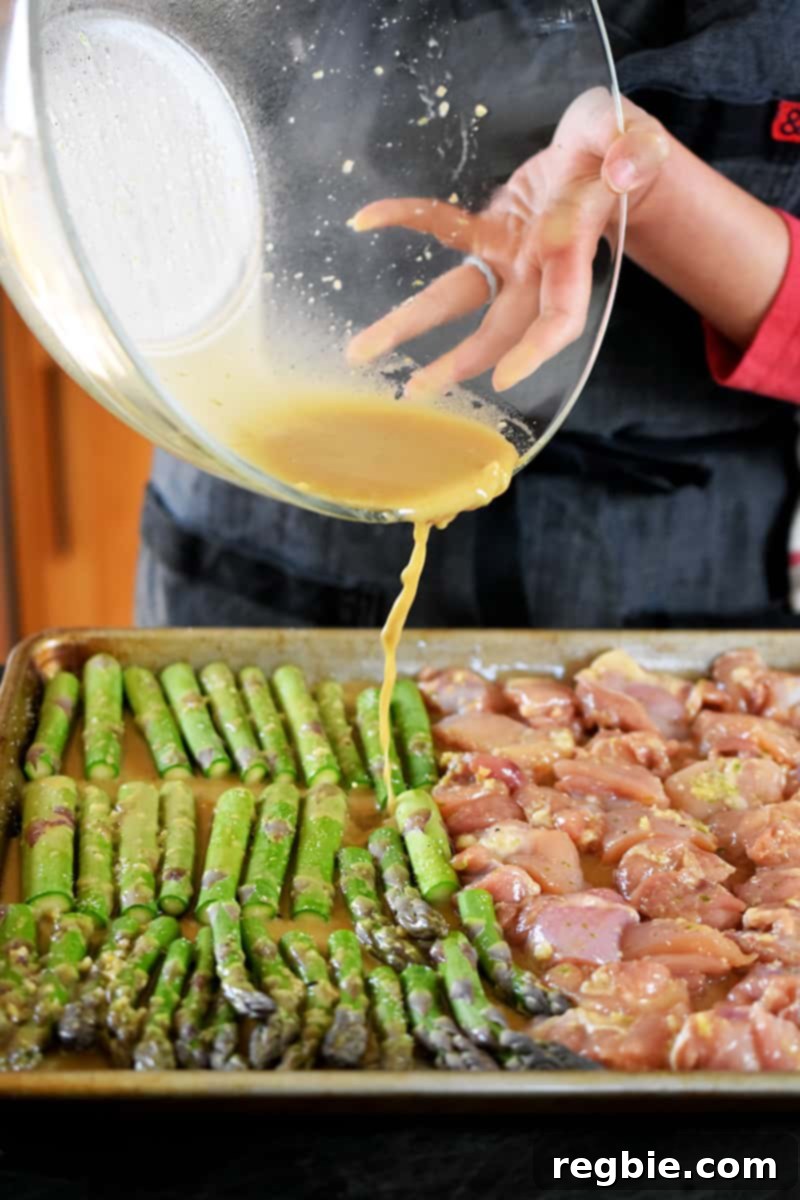 Simple Sheet Pan Chicken & Asparagus: Paleo Perfection 13 A rich, glossy marinade is being poured from a large glass bowl onto a sheet pan already filled with perfectly cubed chicken and fresh asparagus, ensuring every ingredient is coated.