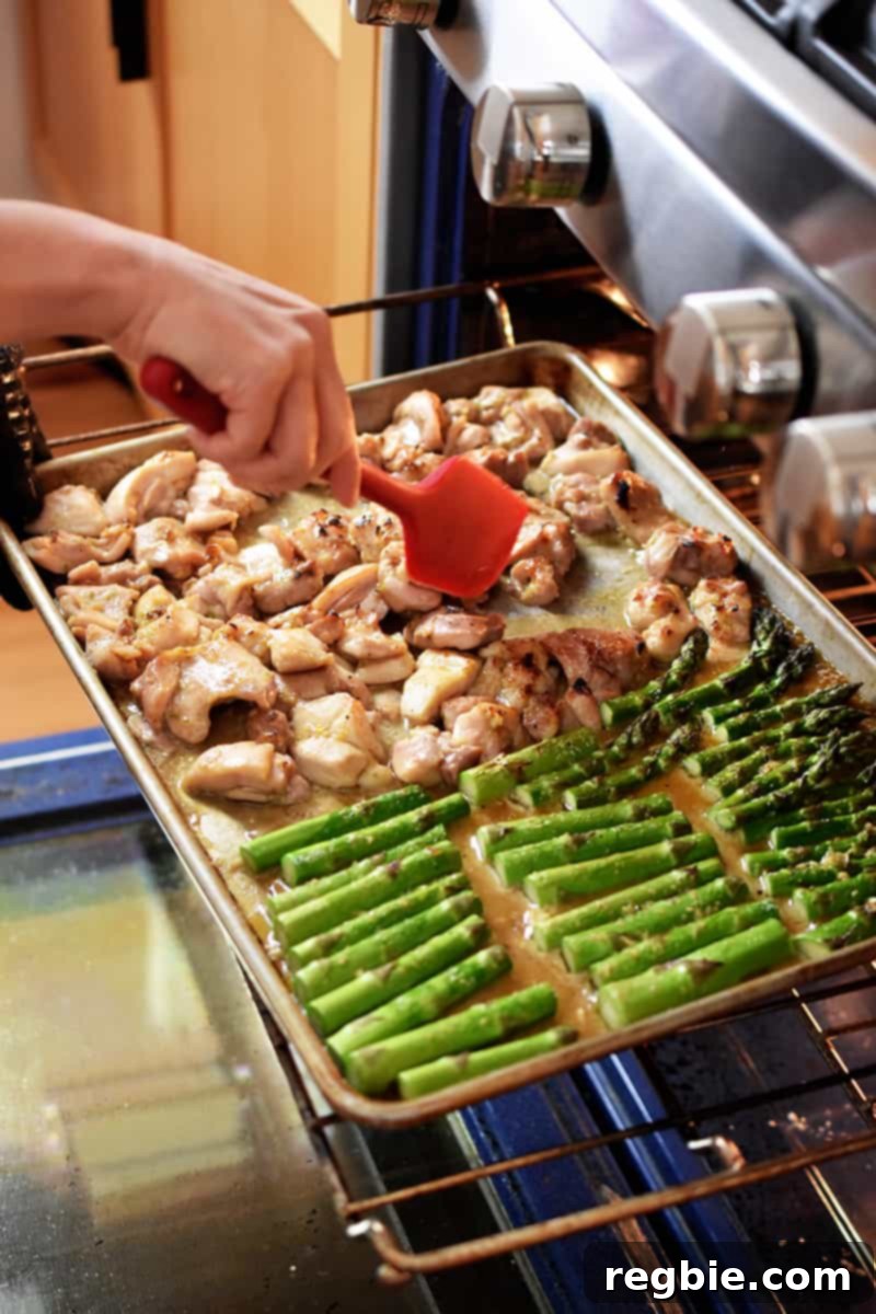 Simple Sheet Pan Chicken & Asparagus: Paleo Perfection 14 Inside an open oven, a person uses a red silicone spatula to gently stir the sheet pan chicken and asparagus, ensuring even cooking as it broils to perfection.
