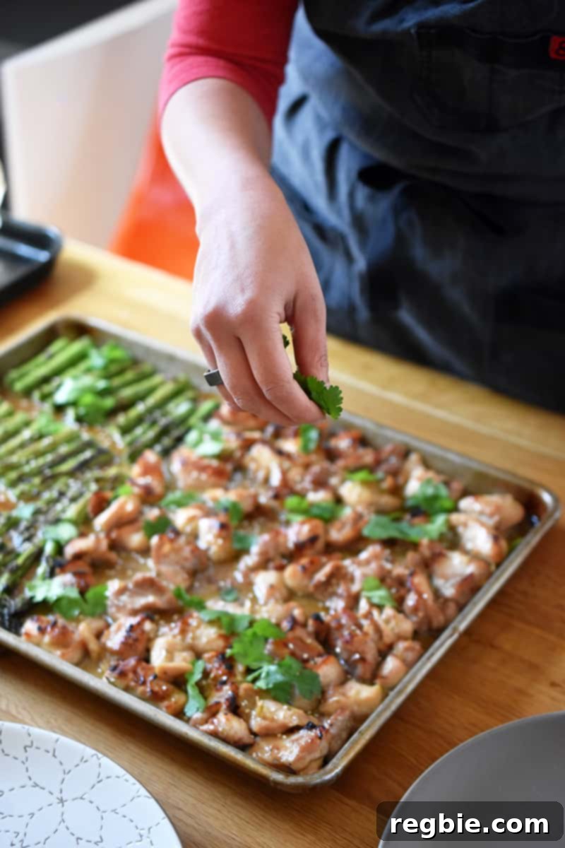 Simple Sheet Pan Chicken & Asparagus: Paleo Perfection 5 A hand is delicately sprinkling vibrant fresh cilantro over a sheet pan generously filled with succulent chicken pieces and tender asparagus spears, adding a final touch of freshness.