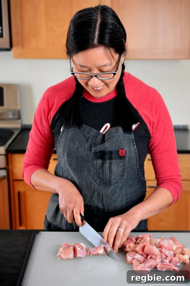 Simple Sheet Pan Chicken & Asparagus: Paleo Perfection 6 An Asian woman, dressed in a gray apron and a red and black shirt, meticulously cuts boneless, skinless chicken thighs into uniform, bite-sized pieces on a cutting board, preparing them for the marinade.