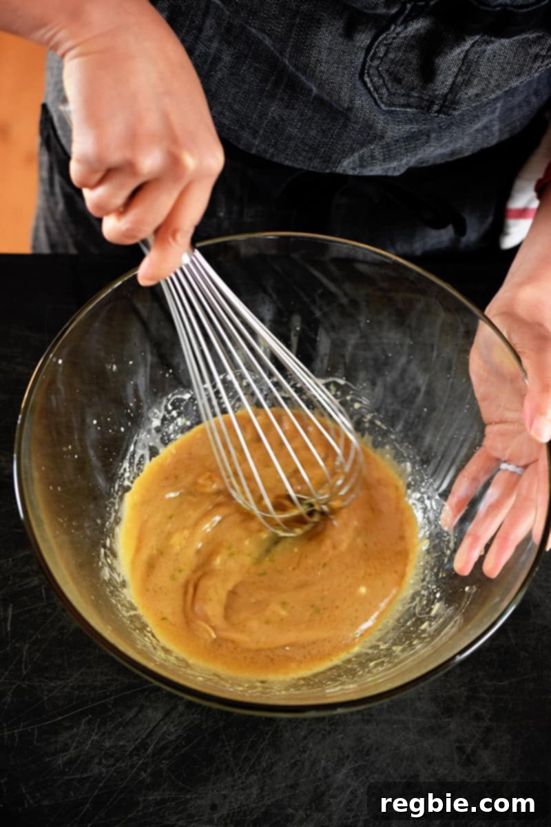 Simple Sheet Pan Chicken & Asparagus: Paleo Perfection 8 An overhead shot captures a person in a gray apron expertly whisking a rich brown sauce in a clear glass bowl, preparing the flavorful marinade for the sheet pan dinner.