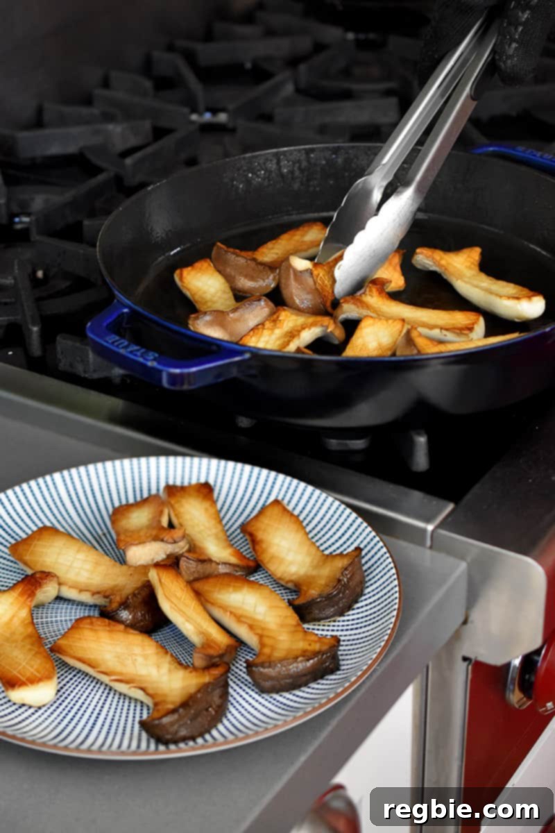 A blue and white plate has golden brown halved king oyster mushrooms and a pair of tongs are transferring the rest from a large frying pan.