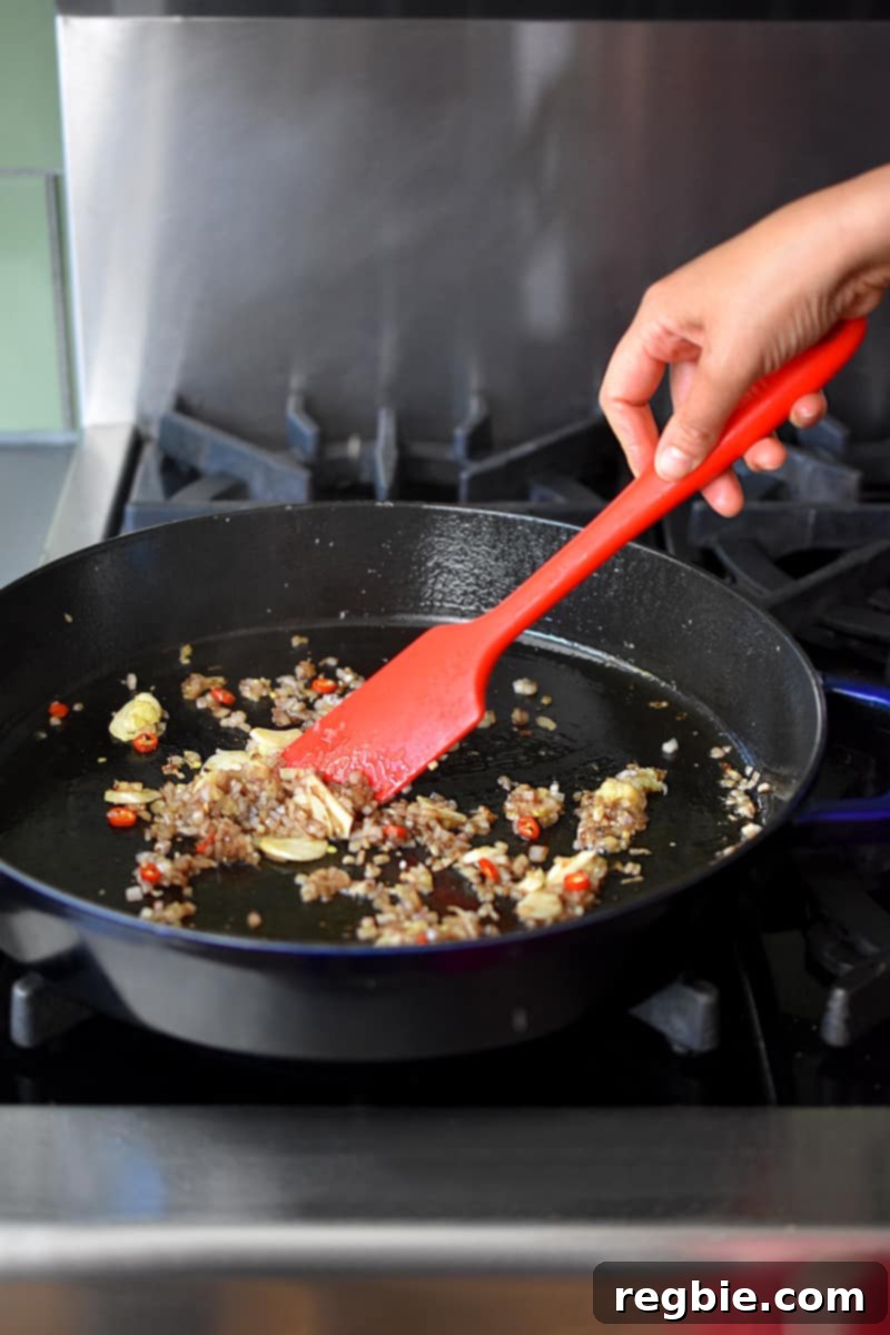 A red spatula is stirring minced shallots, ginger, and garlic in a large cast iron skillet.