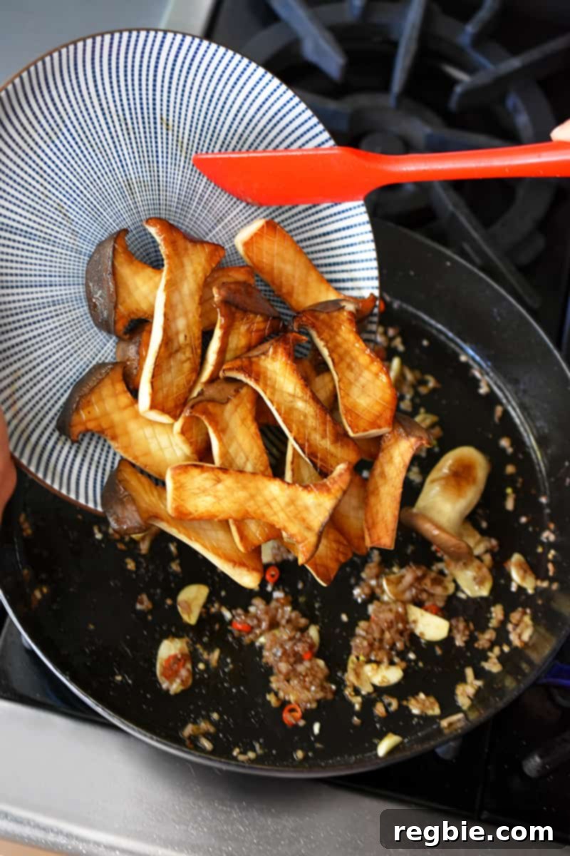 A blue and white plate filled with seared king oyster mushrooms is being added to a cast iron skillet with sautéed diced shallots, ginger, garlic, and Thai Chili peppers