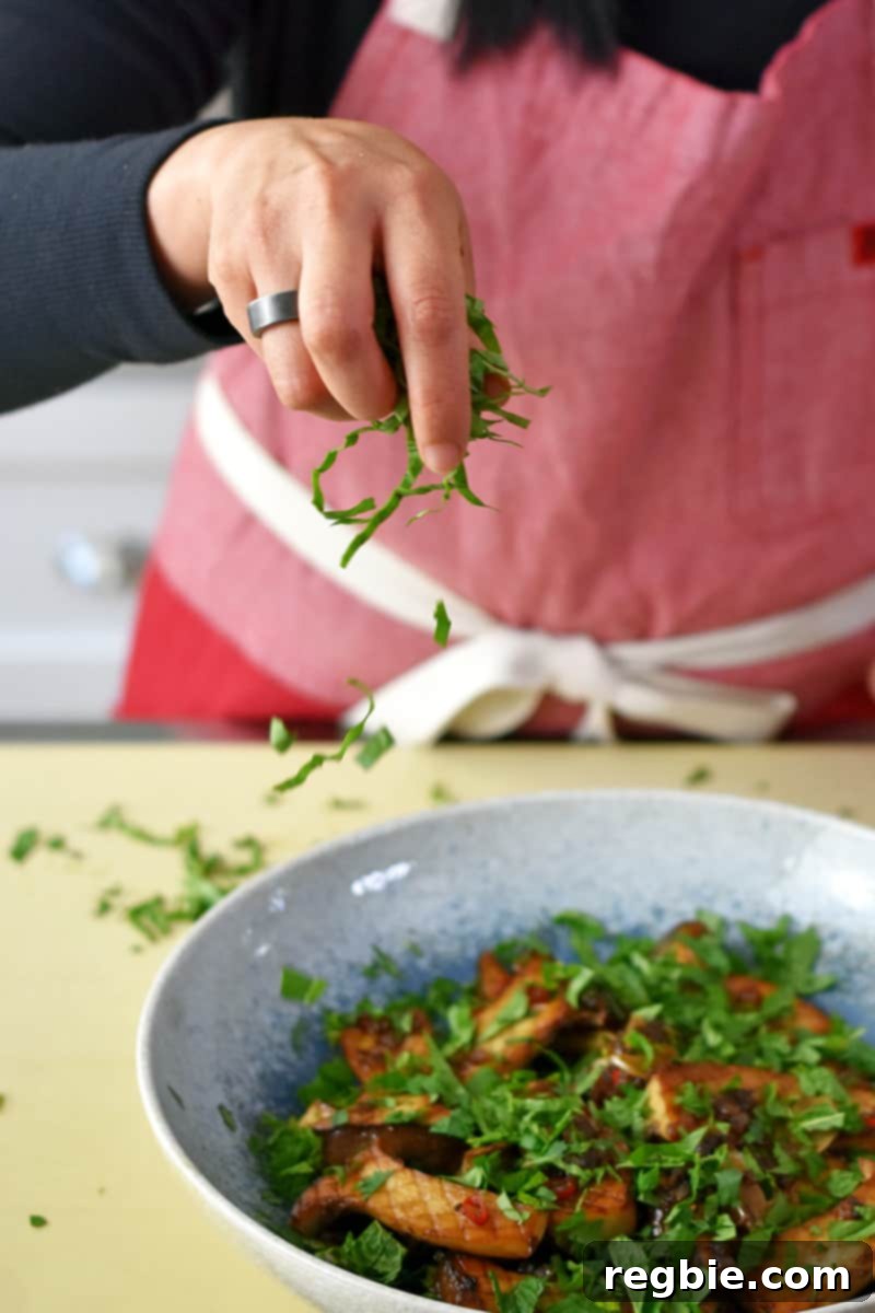 Someone in a red apron is sprinkling minced Thai basil, cilantro, and mint to a bowl filled with pan-seared king oyster mushrooms.
