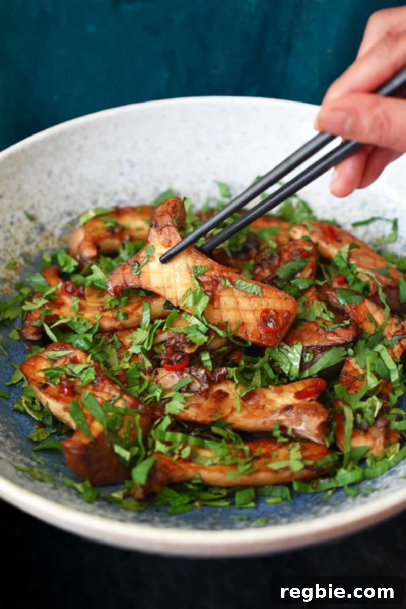 A pair of black chopsticks is picking up a piece of King trumpet mushrooms with Thai Chili peppers and lime from a light blue bowl.