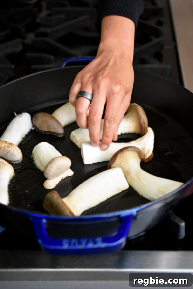 A hand is placing halved king oyster mushrooms cut-side down in a large cast iron skillet