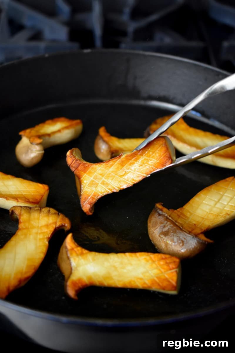 A pair of tongs is flipping golden brown halved king oyster mushrooms in a cast iron pan.