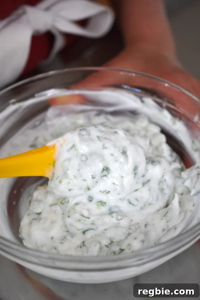 A closeup of the herby coconut yogurt being mixed in a clear glass bowl with a small yellow spatula.