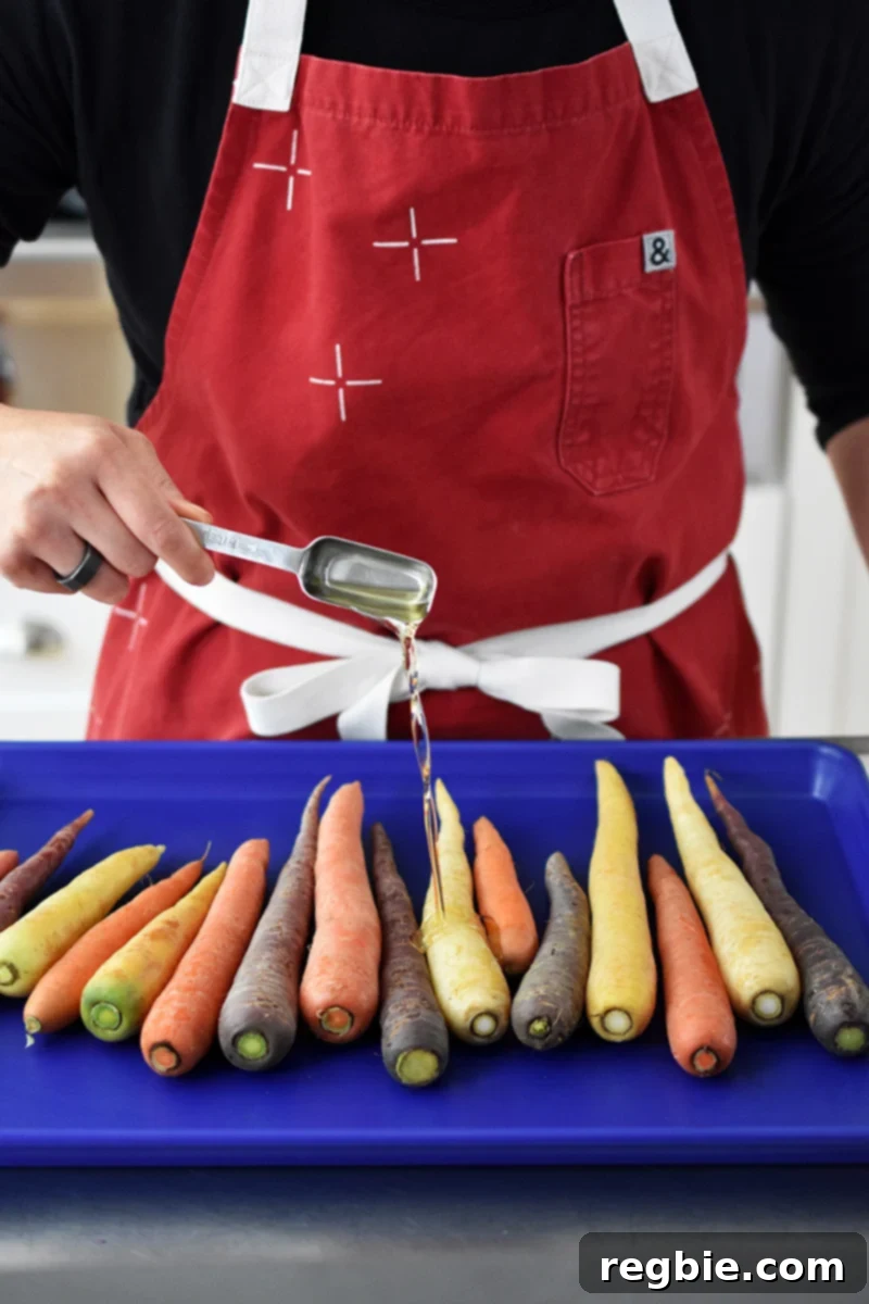 A person in a red apron is drizzling avocado oil on a rimmed baking sheet filled with colorful carrots.