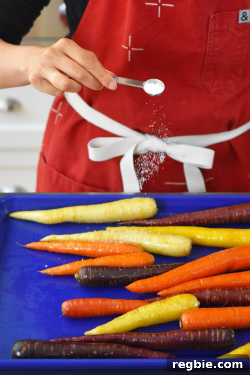 A person in a red apron is sprinkling salt on a blue rimmed baking sheet filled with colorful carrots