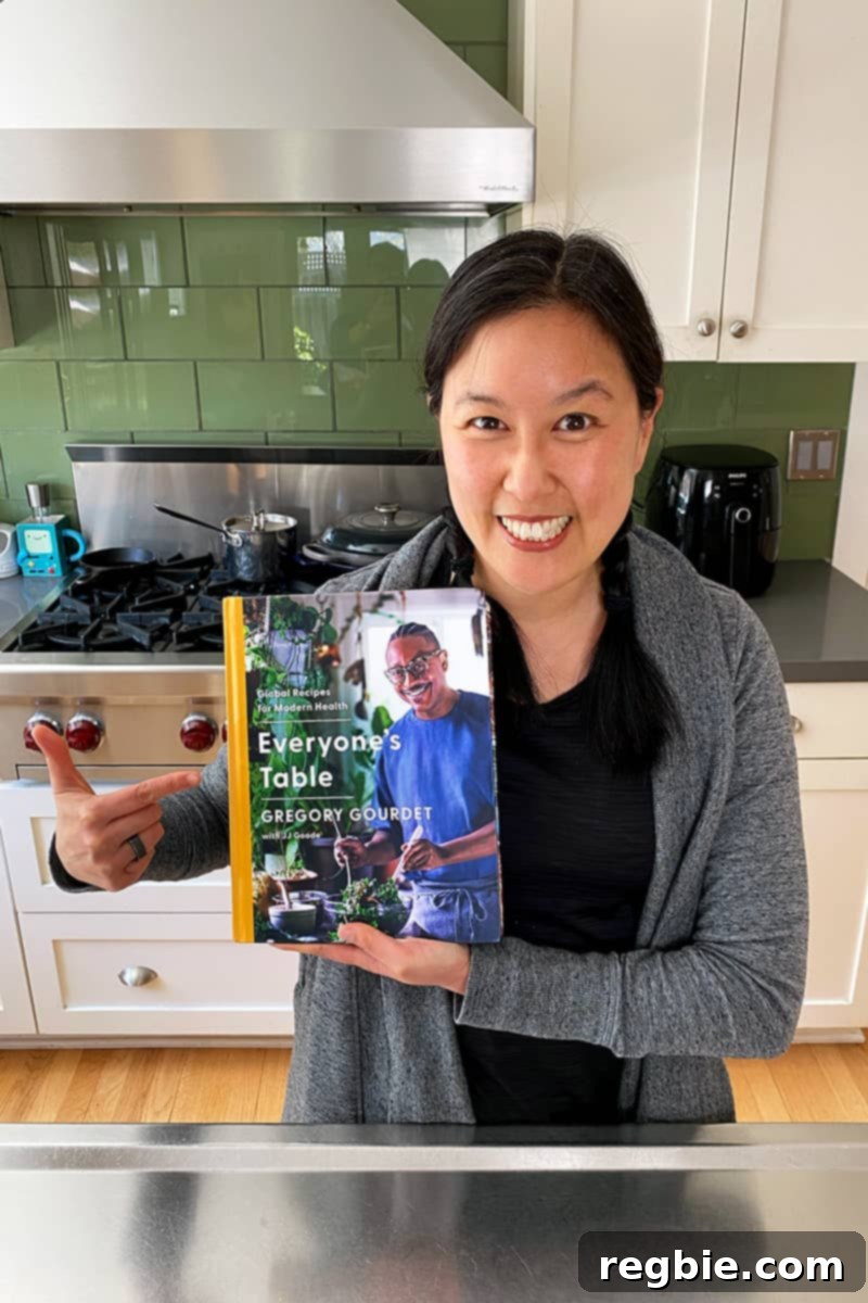 A smiling Asian woman in a kitchen is holding up a copy of Gregory Gourdet’s Everyone's Table cookbook