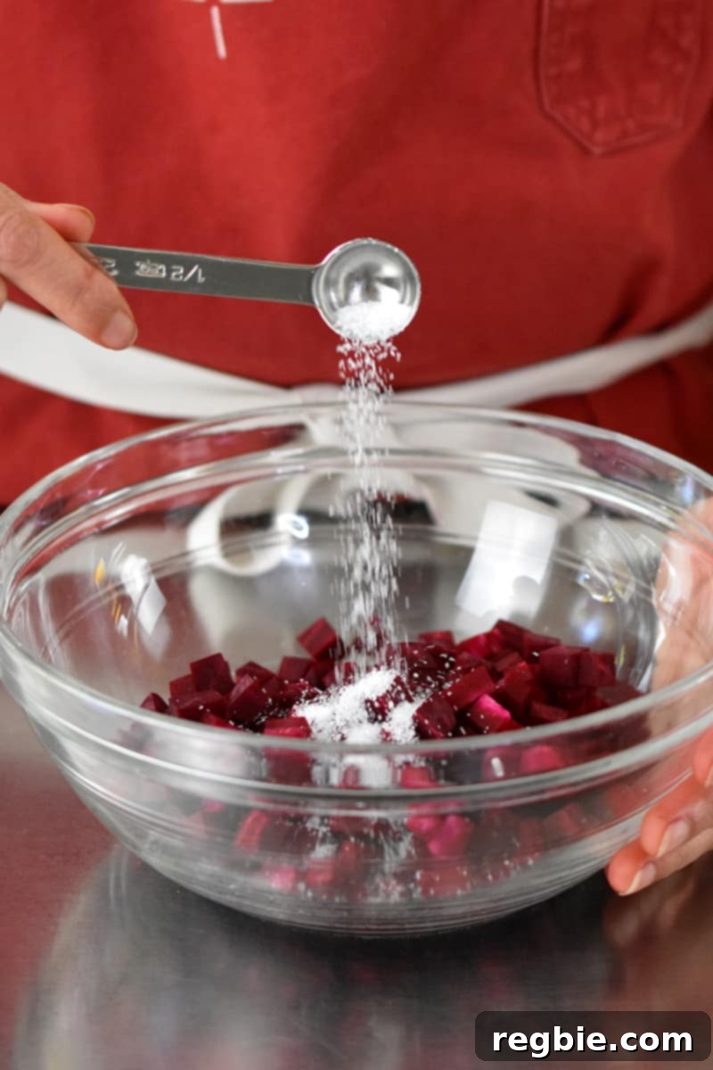 Someone in a red apron is adding a spoonful of salt to a clear bowl filled with diced purple beets