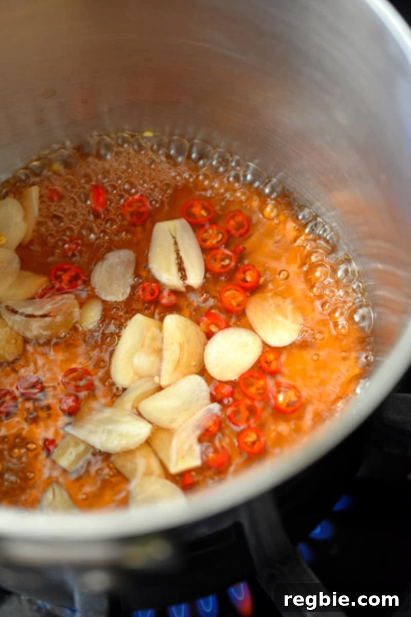 An overhead shot of a small saucepan filled with simmering vinegar, Thai chili peppers, and sliced garlic.