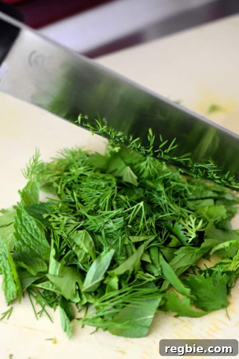 An overhead shot of a chef's knife cutting a mixture of herbs on a beige cutting board.