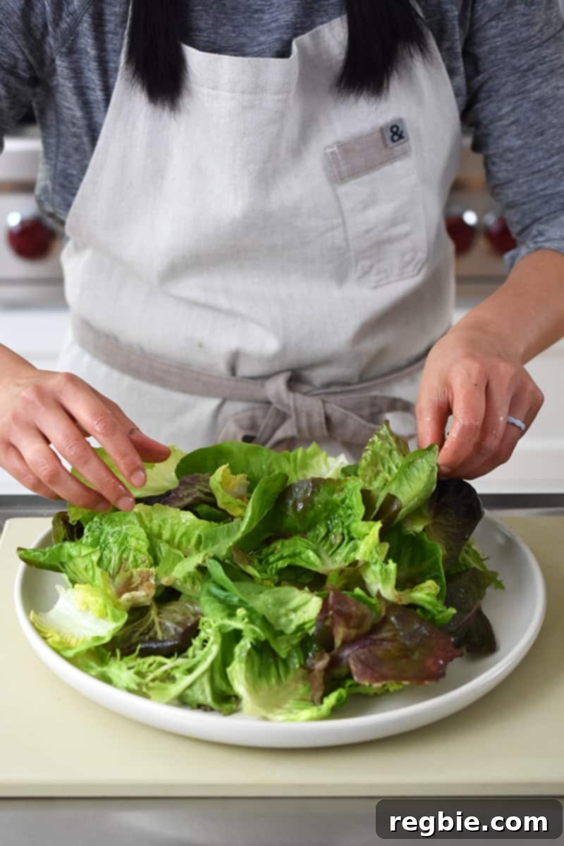 Irresistible Umami Avocado Salad with Garlic Anchovy Vinaigrette 12 A person in a white apron is arranging mixed baby lettuce on a platter.