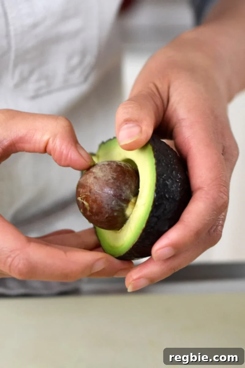 Irresistible Umami Avocado Salad with Garlic Anchovy Vinaigrette 13 A person in a white apron is pulling an avocado pit from a halved avocado