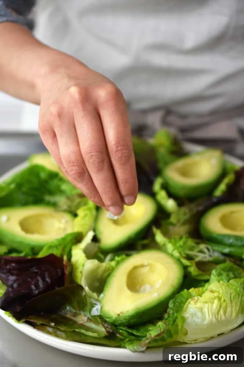 Irresistible Umami Avocado Salad with Garlic Anchovy Vinaigrette 14 A person in a white apron is sprinkling salt on an avocado salad