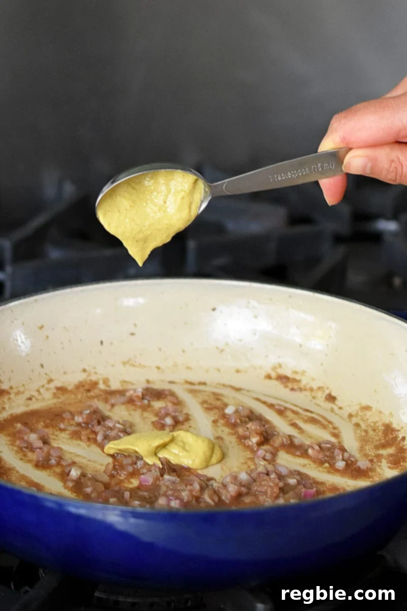 Irresistible Umami Avocado Salad with Garlic Anchovy Vinaigrette 10 A hand is adding a measuring spoon filled with Dijon mustard into a skillet filled with garlic anchovy vinaigrette.