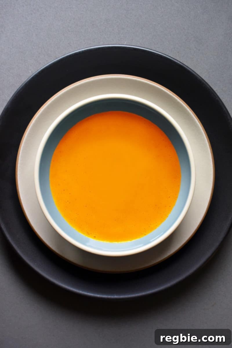 An overhead shot of carrot ginger soup with cardamom in a shallow soup bowl, garnished with fresh herbs.