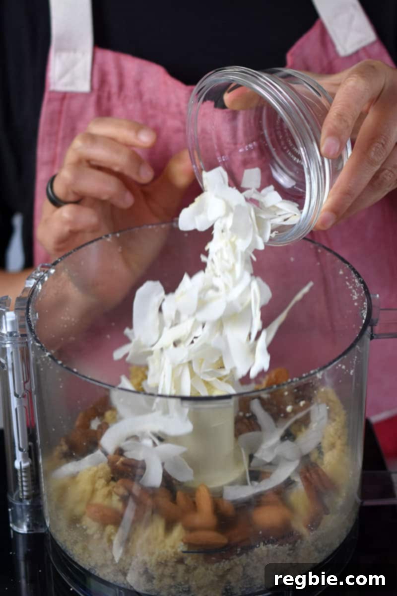 A person in a red apron is adding coconut flakes to an open food processor to make a paleo berry crisp.