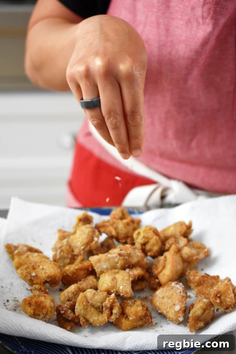 A person in a red apron is sprinkling crushed Sichuan peppercorns and salt on gluten-free Taiwanese popcorn chicken.