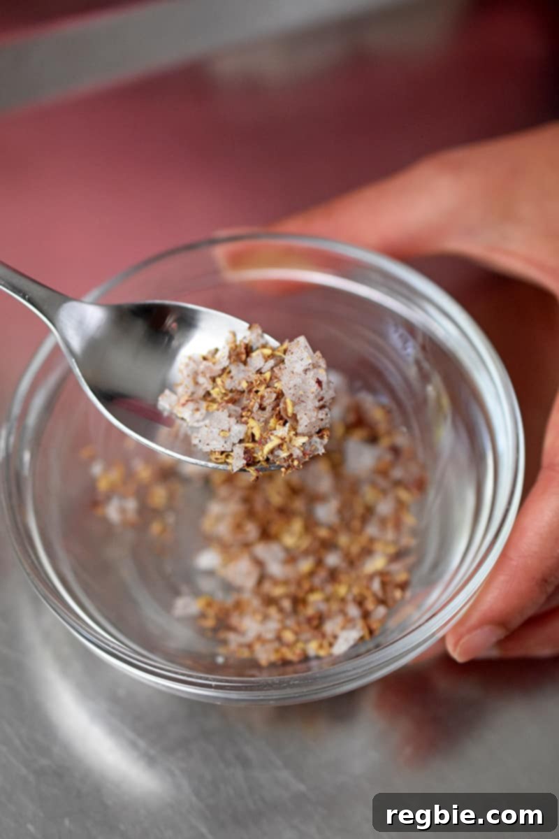 An overhead shot of a spoon mixing crushed Sichuan pepper corns with flaky sea salt in a small bowl.