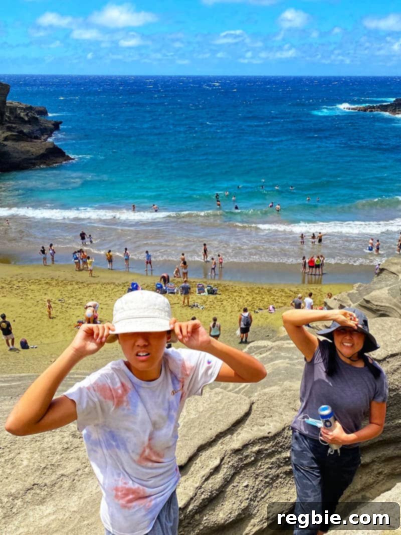 A teenage boy and an Asian woman are holding their hats to their head while they stand on a beautiful but windy beach on the Big Island.