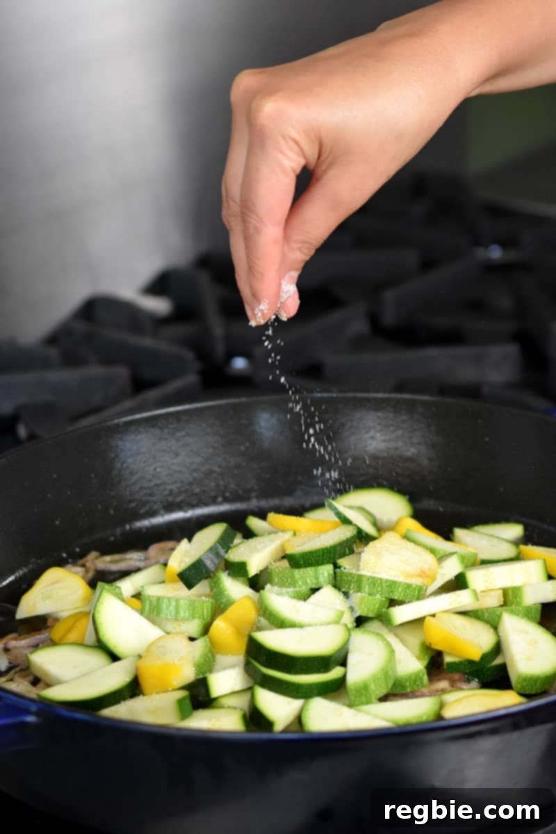 A hand sprinkling salt onto sliced summer squash in a cast iron skillet