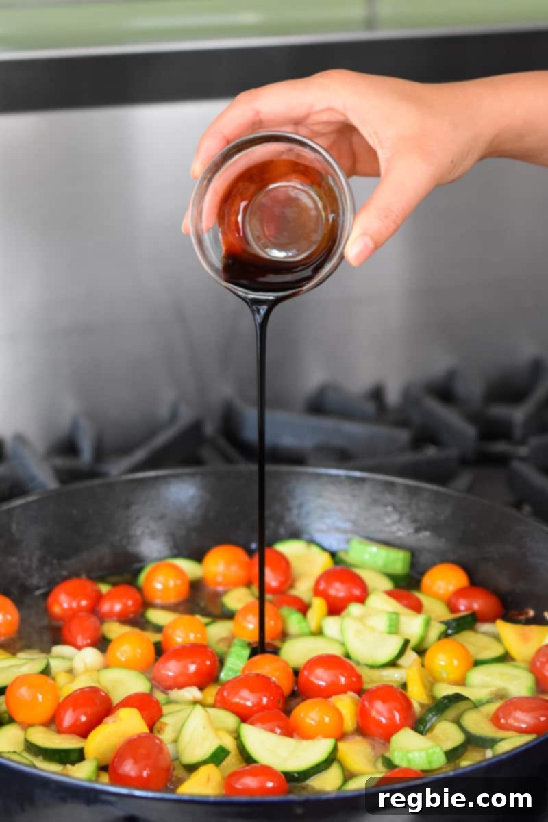 A hand is pouring aged balsamic vinegar into a pan filled with sautéed summer squash and cherry tomatoes