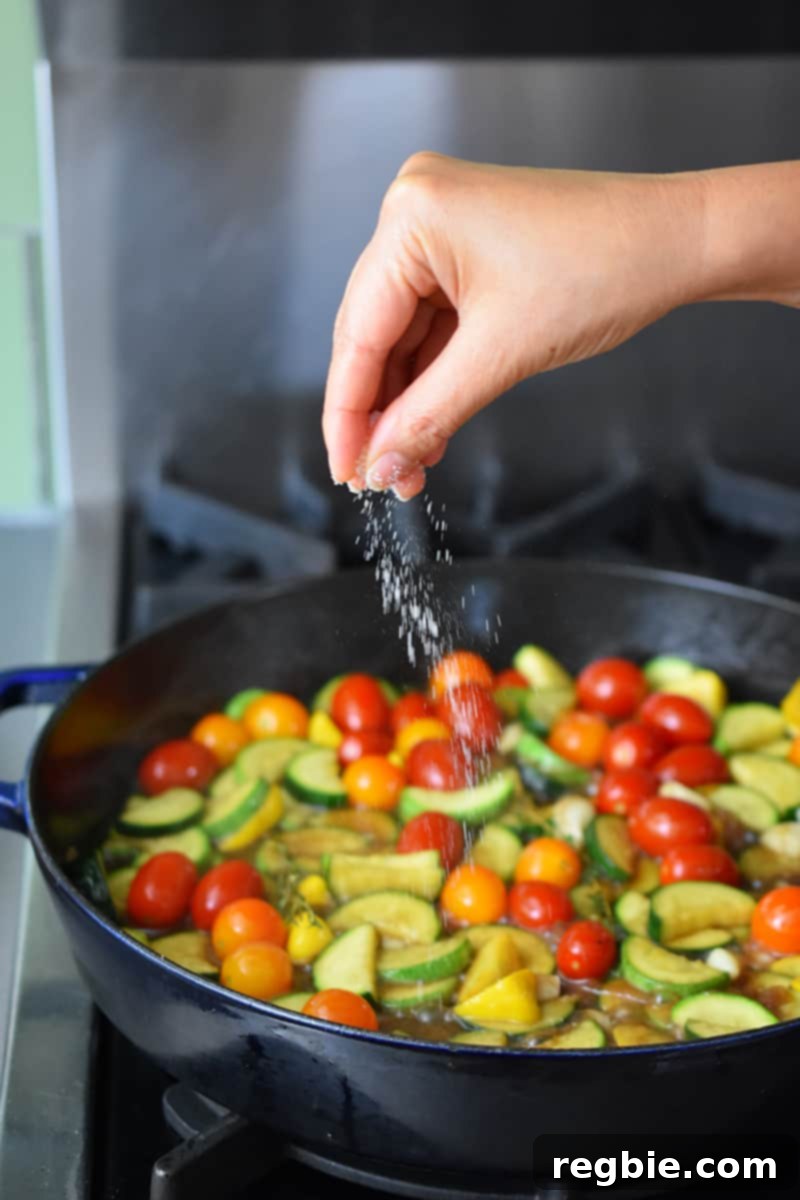 A hand is adding salt to a skillet filled with sliced summer squash, cherry tomatoes, garlic, shallot, and thyme.
