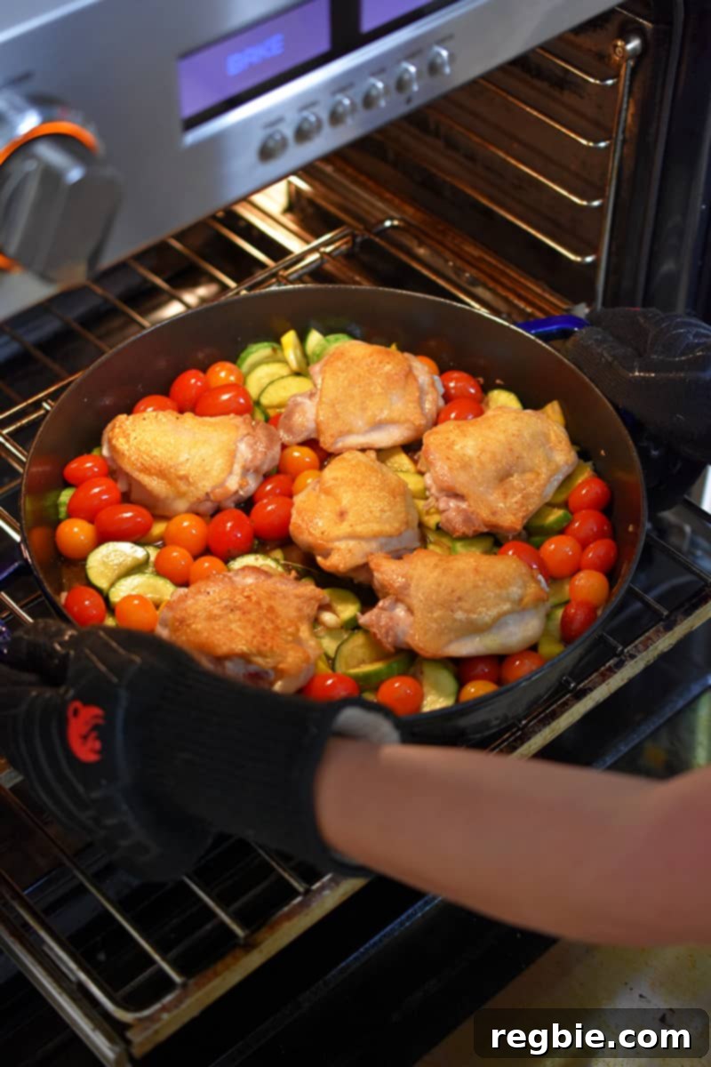 Someone placing a skillet with crispy chicken thighs and summer vegetables into an open oven