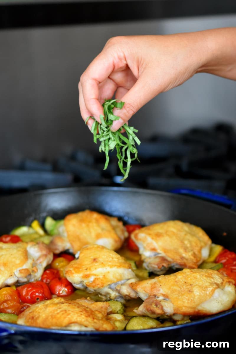 A hand is adding finely sliced fresh basil to a skillet of crispy baked chicken thighs and summer vegetables