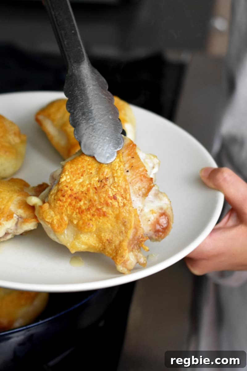 A pair of tongs transferring crispy pan seared chicken thighs on a cream colored plate.