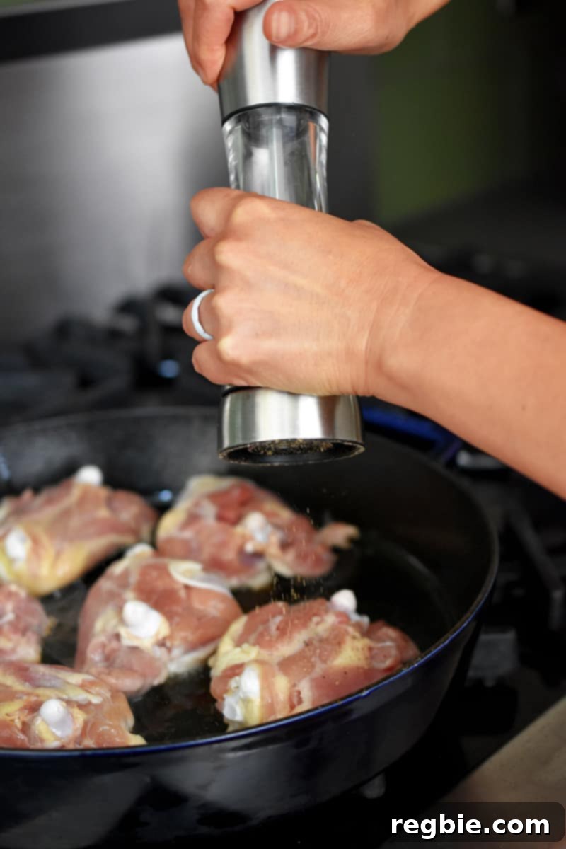 Someone adding freshly cracked black pepper from a pepper mill to the meaty side of chicken thighs in a frying pan.