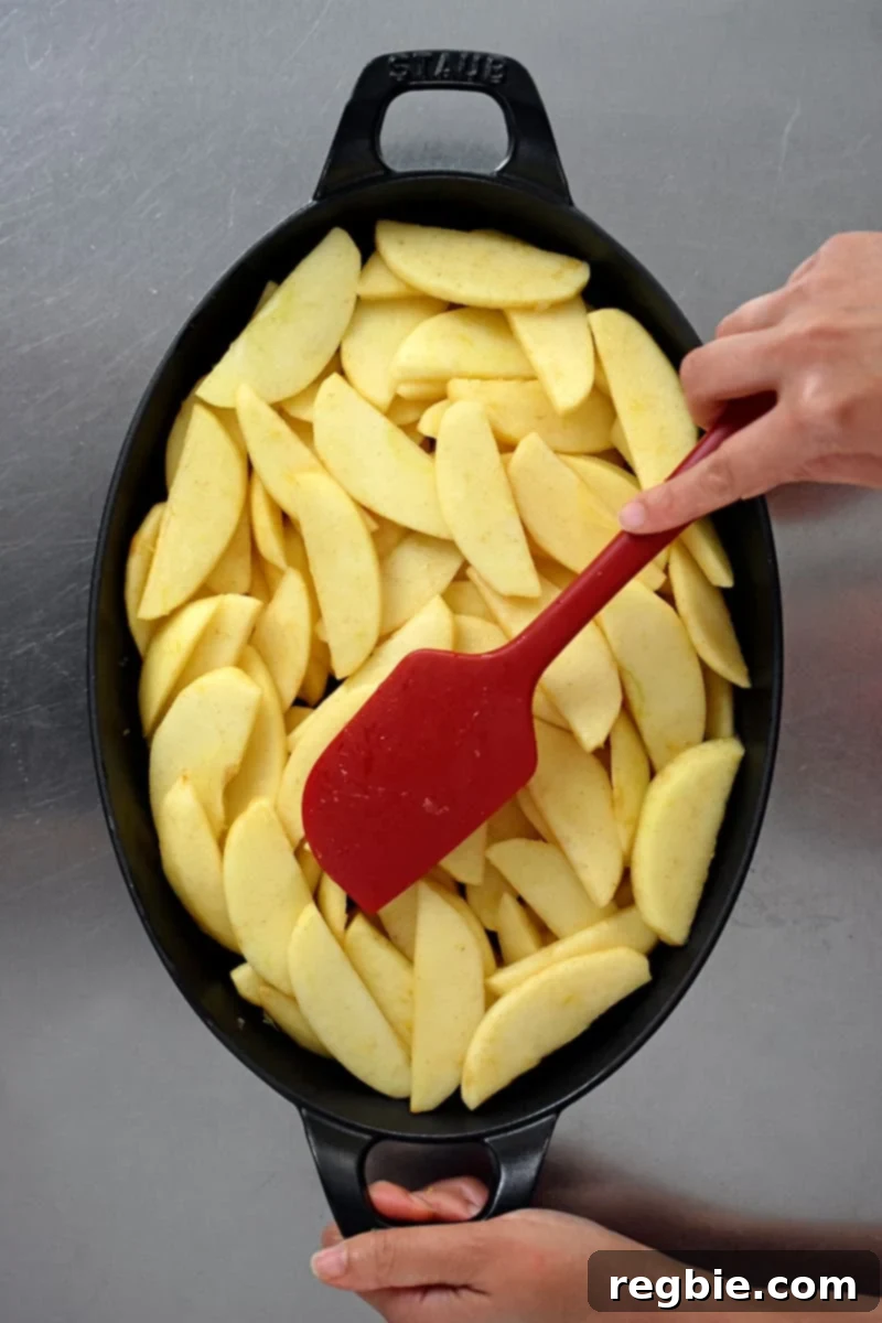Guilt Free Apple Crisp 16 An overhead shot of an oval baking dish filled with sliced apples. A red silicone spatula is making an even layer.