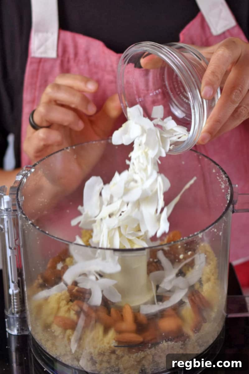 Guilt Free Apple Crisp 10 A person in a red apron is adding coconut flakes to a food processor filled with paleo apple crisp topping ingredients.