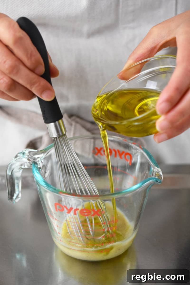 A small bowl of extra virgin olive oil is being drizzled into a measuring cup that is filled with champagne vinaigrette ingredients being whisked.