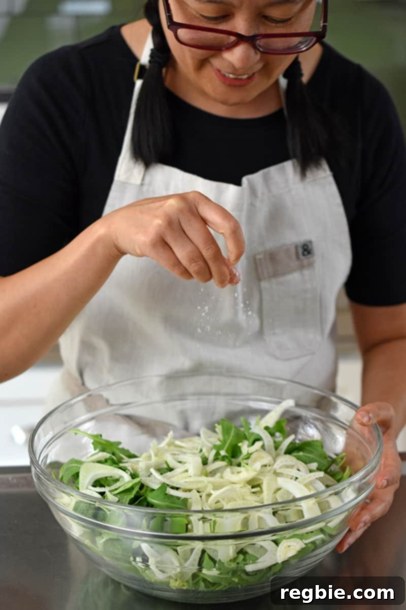 An Asian woman in glasses is seasoning some baby arugula and shaved fennel in a large bowl.